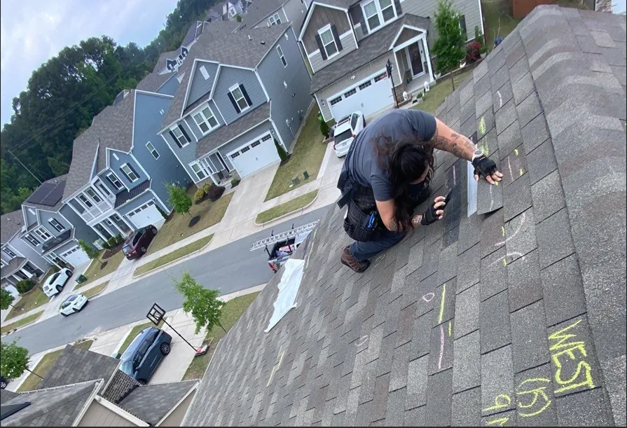 Roofer kneeling on a shingle roof marked with chalk during a damage inspection